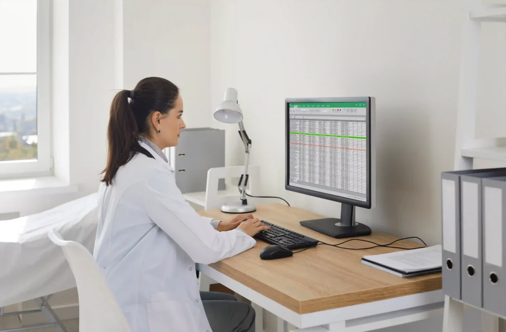 Woman in a white lab coat seated at a desk, typing on a keyboard while reviewing a spreadsheet on a computer monitor.